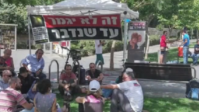 A group of people gathered outdoors, some seated, with a large banner in the background.