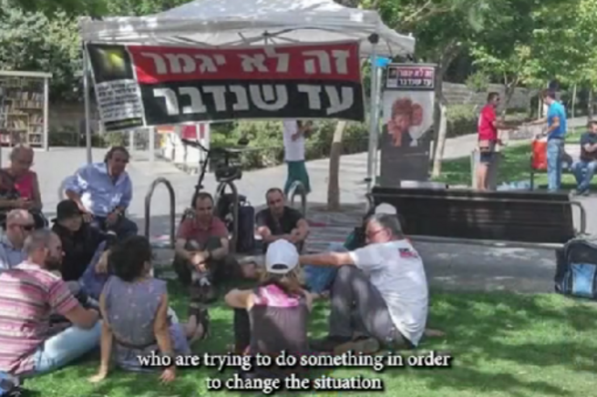 A group of people gathered outdoors, some seated, with a large banner in the background.