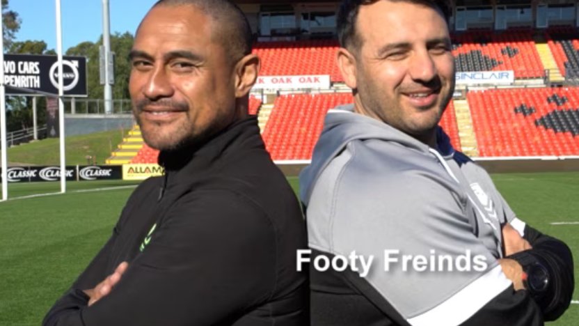 Two men smiling at a stadium, one with arms crossed.