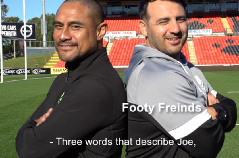 Two men smiling at a stadium, one with arms crossed.